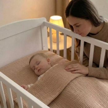 Muślinowa pościel do łóżeczka niemowlęcego w odcieniu ciemny beż. Śpiące dziecko i czuwająca obok mama